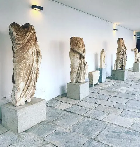 Mikonos Arkeoloji Müzesi'nin beyaz duvarlı, taş zeminli bir galerisinde, çeşitli yükseklerdeki kaideler üzerinde sergilenen, başsız ve kolsuz, antik dönemlere ait, dökümlü kıyafetli taş heykeller, Ege'nin zengin tarihini yansıtıyor.