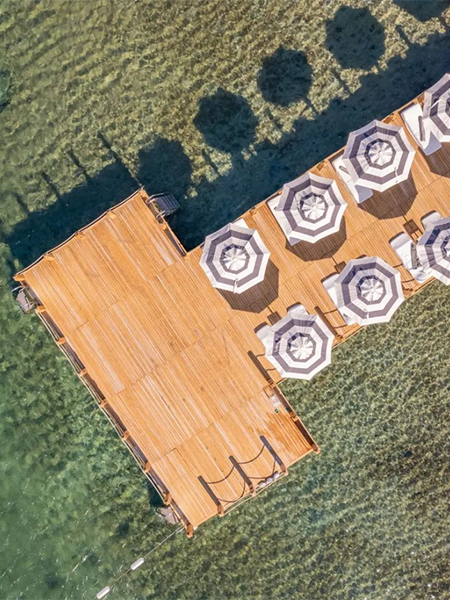 Bodrum'un en iyi otellerinden birinde lüks bir tatil vadeden, berrak turkuaz sulara uzanan ahşap iskelede, beyaz şezlongların üzerini gölgeleyen siyah beyaz çizgili şemsiyeler net gölgeleriyle huzurlu bir atmosfer yaratıyor.