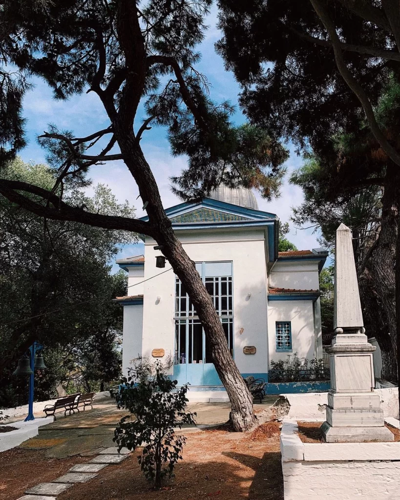 Ön planda kalın bir ağaç gövdesinin kısmen kapattığı, mavi kubbeli beyaz bir yapı olan Aya Yorgi Kilisesi, taş patikaların, bankların ve çanların bulunduğu yemyeşil bir bahçe ile çevrili olup, tarihi atmosferi ve açık mavi gökyüzüyle dikkat çekiyor.
