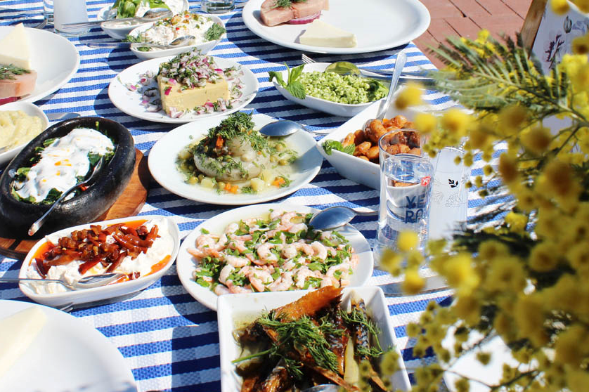 Mavi beyaz çizgili bir masa örtüsü serili, güneşli bir sahil restoranı ortamında; taze karides salatası, enginar, fava, yoğurtlu meze ve kadehlerde rakı gibi bol çeşitli Türk mezeleriyle donatılmış, iştah açıcı bir öğle yemeği sofrası yakın çekim olarak görülüyor.