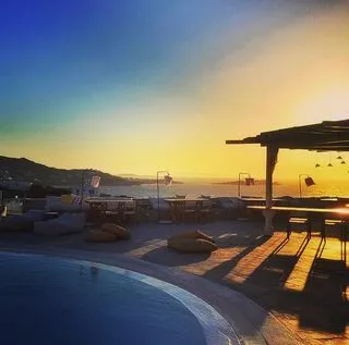 Mikonos'un tepelerinden Ege Denizi'ne nazır bir konumda, gün batımının altın renklerine bürünmüş gökyüzü altında, lüks bir otelin sonsuzluk havuzu ve ahşap çatılı bohem tarzı oturma alanları, keyifli anlar için ideal bir eğlence noktası olarak ziyaretçileri bekliyor.