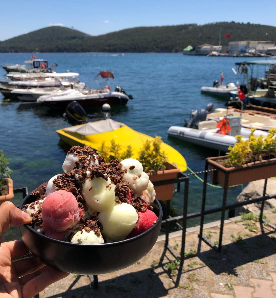 Güneşli bir yaz gününde, mavi denize açılan tekneler ve yemyeşil adanın eşlik ettiği sakin bir iskele manzarasında, bir elin tuttuğu siyah kasede pembe, sarı ve beyaz dondurma toplarının bolca granül ile süslenmiş iştah açıcı görünümü.