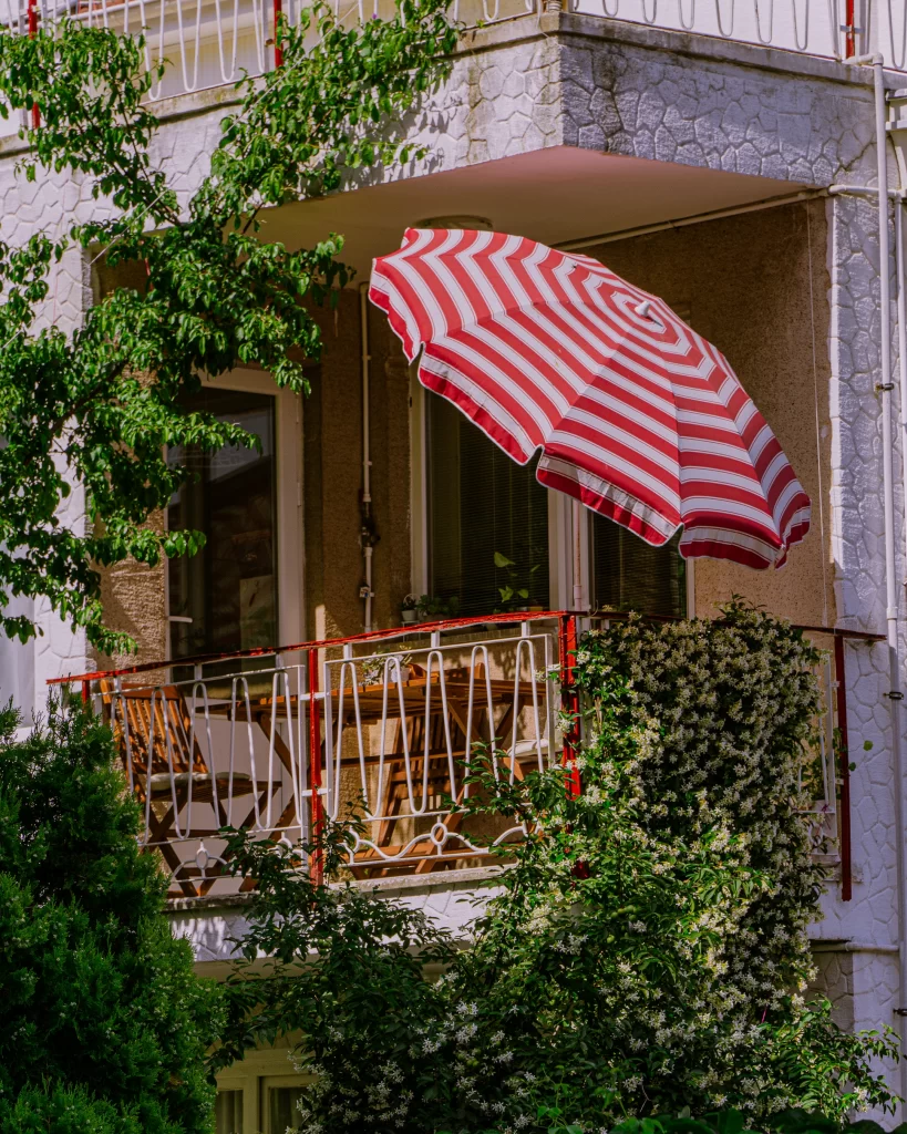 Yeşil bitkilerle sarılmış, kırmızı beyaz çizgili bir şemsiyenin gölgesindeki ahşap masa ve sandalyelerin bulunduğu sakin bir balkon, Burgazada'ya gidilecek en güzel zamanları çağrıştıran huzurlu bir atmosfer sunuyor.
