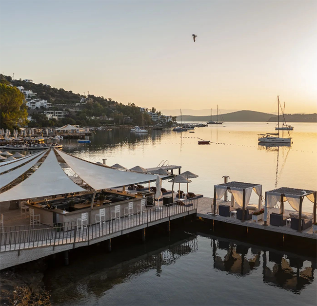 Bodrum'un en iyi otellerinden birinde gün batımının yansıdığı sakin Ege suları üzerinde, beyaz tenteli modern bir bar alanı ve şık kabinlerin yer aldığı uzun ahşap iskelede tekneler eşliğinde huzurlu bir akşamüstü manzarası.