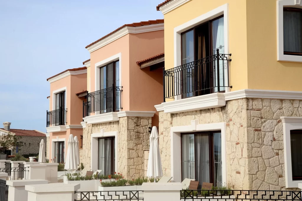 Sıra sıra dizilmiş, pastel sarı ve şeftali rengi cephelere sahip, alt katları taş kaplamalı, demir balkon korkulukları ve teraslarında beyaz katlanmış şemsiyeleri olan modern villalar mavi gökyüzü altında güneşli bir günde yer alıyor.