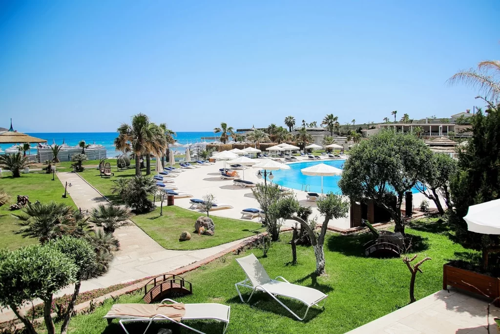Güneşli bir günde, masmavi denizin kıyısında uzanan yemyeşil bir tatil köyünde, berrak mavi yüzme havuzu, çevresindeki şık beyaz şezlonglar, şemsiyeler ve palmiye ile küçük ağaçlarla süslenmiş yollar dinlendirici bir tatil atmosferi oluşturuyor.