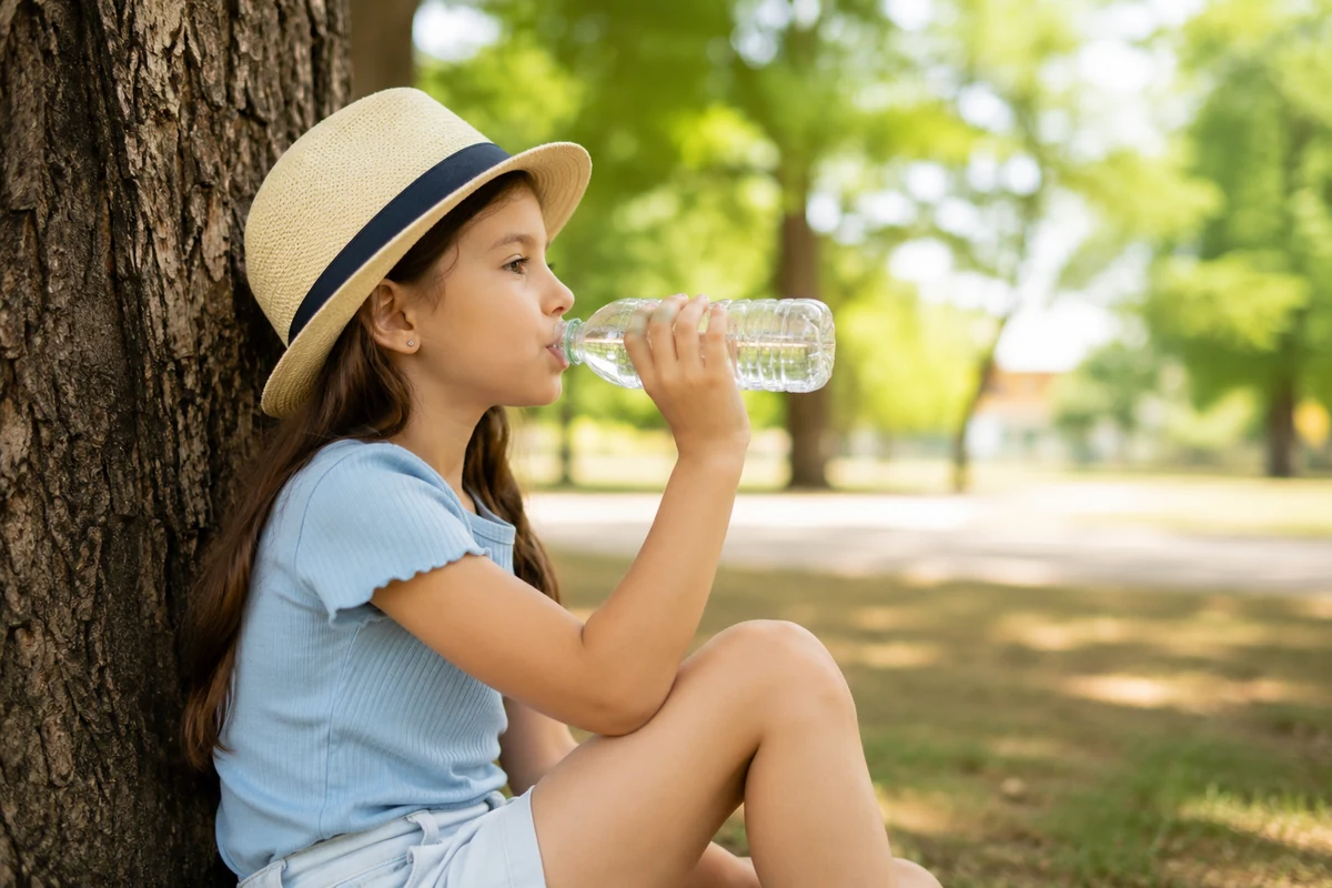 Hasır şapkasıyla bir ağacın gölgesine yaslanmış, ışıklı yeşil park zemininde oturan genç bir kız, elindeki plastik şişeden büyük bir yudum su içerek çocuklarda su tüketiminin ne kadar önemli olduğunu doğal ve huzurlu bir şekilde gözler önüne seriyor.