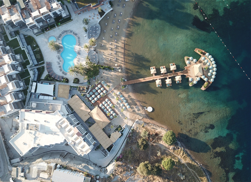 Bodrum'un en iyi otellerinden biri olan D'Plaj Bodrum Yalıkavak'ı kuşbakışı gösteren bu geniş planda, turkuaz Ege sularına uzanan ahşap bir iskele, üzerinde şemsiyelerle donatılmış lüks dinlenme locaları, bembeyaz kumlu özel plajı, palmiye ağaçları ile çevrili büyük havuz alanı ve çevresindeki modern konaklama binaları dikkat çekiyor.