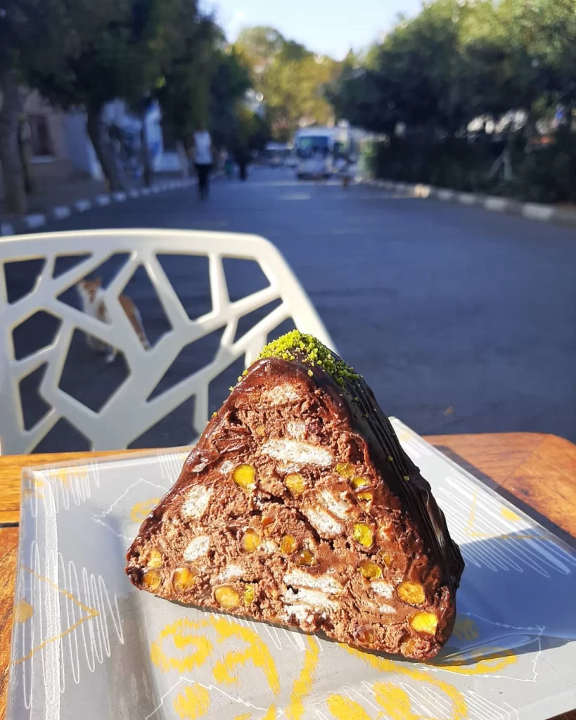 Dekoratif desenli bir tabakta, üstü çikolata soslu ve ince çekilmiş fıstıklarla süslenmiş, bol bisküvi ve Antep fıstığı parçacıkları içeren, üçgen şeklinde iştah açıcı bir mozaik pasta dilimi; arka planda ağaçlıklı bir sokak, bir sandalye ve bir kediyle dış mekanda güneşli bir öğle anını betimliyor.