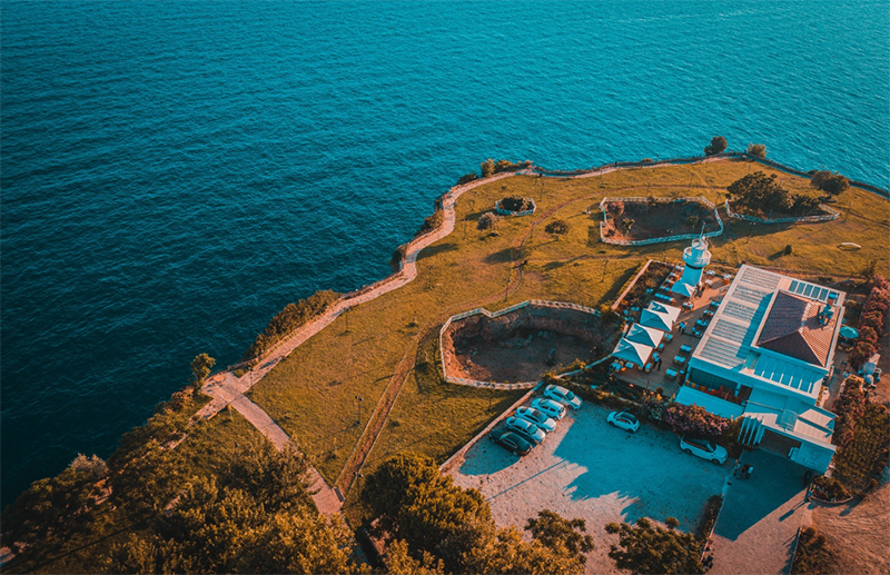 Antalya'nın en iyi restoranlarından Fener Bababurnu, turkuaz Akdeniz'in kıyısındaki yemyeşil bir falez üzerinde, açık hava oturma alanları, beyaz bir deniz feneri ve otoparkı ile çevrili modern binalarıyla kuşbakışı görünüyor, ziyaretçilerine eşsiz bir manzara sunuyor.