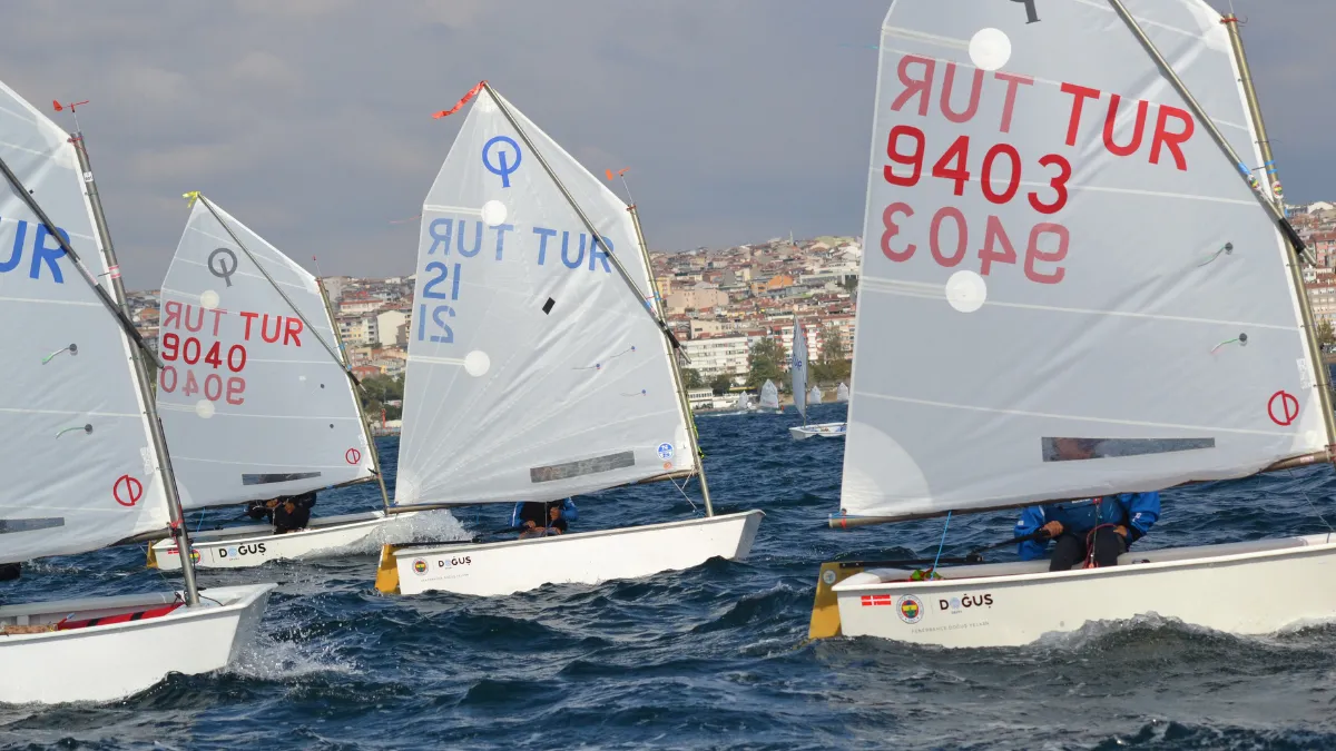Fenerbahçe Yelken Kulübü logolarının belirgin olduğu teknelerde genç sporcuların dalgalı denizde kıyasıya yarıştığı, her bir yelkeninde TUR ibaresi ve numaraların (örneğin 9403, 9040) görüldüğü, arkasında şehir manzarası olan heyecanlı bir yelken yarışı anı.