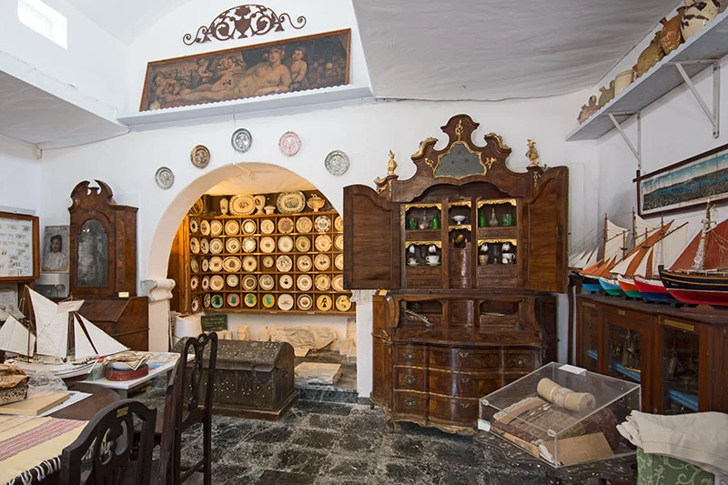 Mikonos Folklor Müzesi'nin içinde, beyaz duvarlar arasında sergilenen geleneksel el yapımı tabaklar, antika ahşap mobilyalar ve renkli yelkenli gemi modelleri, adanın zengin yerel kültürünü bir Mikonos seyahati rehberi için güzelce yansıtıyor.