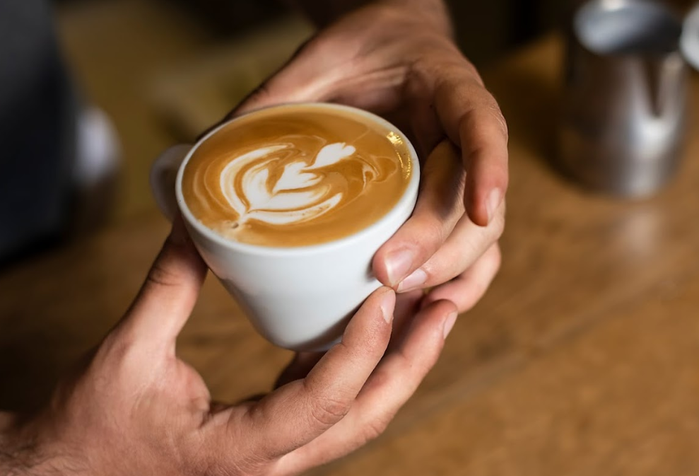 Bir barista veya kahve severin elleri, üzerinde ustalıkla oluşturulmuş yaprak ve kalp desenli kremalı latte artına sahip beyaz bir fincanı zarifçe tutuyor; arka planda hafifçe bulanık ahşap tezgah ve metal sütlük detayları, sıcak bir kahve keyfini çağrıştırıyor.
