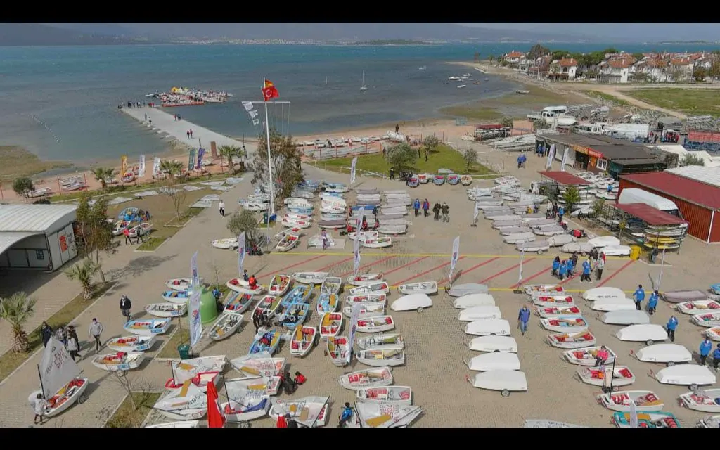 Göztepe Yelken Kulübü tesislerinde, kıyıda onlarca küçük optimist yelkenlinin sıralandığı, sporcuların ve kulüp üyelerinin yoğun bir etkinliğe hazırlandığı veya katıldığı, denize uzanan iskelede de kalabalığın olduğu canlı bir kuşbakışı görünüm.