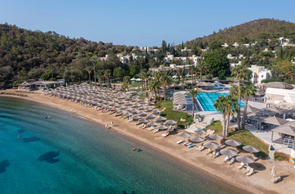 Bodrum'un masmavi koylarından Yalıçiftlik'te bulunan Hapimag Sea Garden Resort'un kuşbakışı görünümünde, uzun kum plajı, sıralı hasır şemsiyeler, palmiye ağaçları ve yeşil tepelere yayılan otel binaları, doğayla iç içe lüks bir Bodrum oteli deneyimini gözler önüne seriyor.