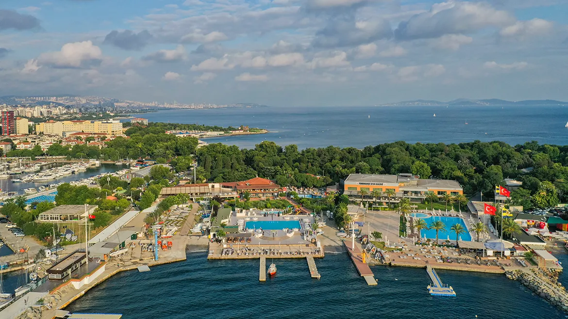 Marmara Denizi kıyısında yer alan İstanbul Yelken Kulübü'nün kuşbakışı manzarasında, yelken ve diğer su sporları için birçok yat ve teknenin demirli olduğu hareketli bir marina, güneşlenen insanların bulunduğu turkuaz renkteki yüzme havuzları, yemyeşil ağaçlık alanlar ve uzak şehir silüeti açıkça görülmektedir.