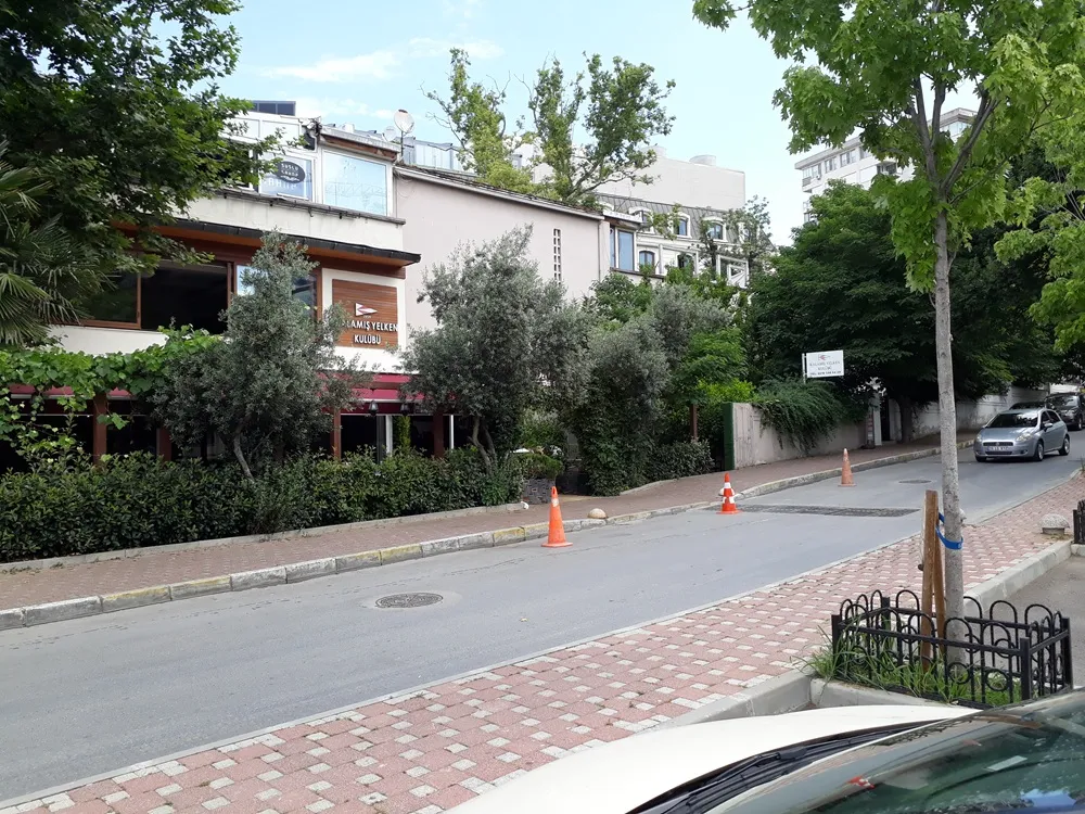 Yeşil ağaçlar ve çalıların çevrelediği, hafif eğimli bir cadde üzerinde yer alan Kalamış Yelken Kulübü'nün ön cephesi, kırmızı tente detayları ve çevresindeki binalarla birlikte gündüz vakti bir görünüm sunuyor.