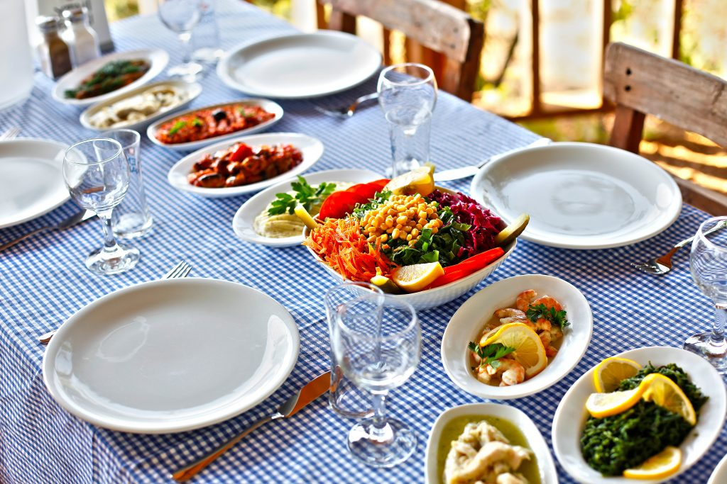 Mavi beyaz kareli bir masa örtüsü serilmiş, açık hava bir restoranda, ortasında nohutlu ve sebzeli renkli bir salata ile karides, ıspanak ve diğer zeytinyağlı mezelerin bulunduğu zengin bir Türk sofrası.