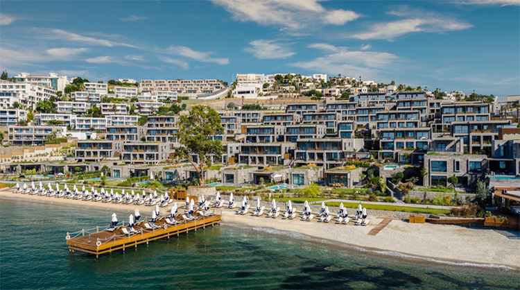 Kaya Palazzo Resort'un modern mimarili binalarının yemyeşil bir yamaca yayıldığı, denize uzanan iskelesi ve altın rengi kumsalıyla lüks bir tatil vaat eden bu Bodrum oteli, mavinin ve yeşilin eşsiz uyumunu gözler önüne seriyor.