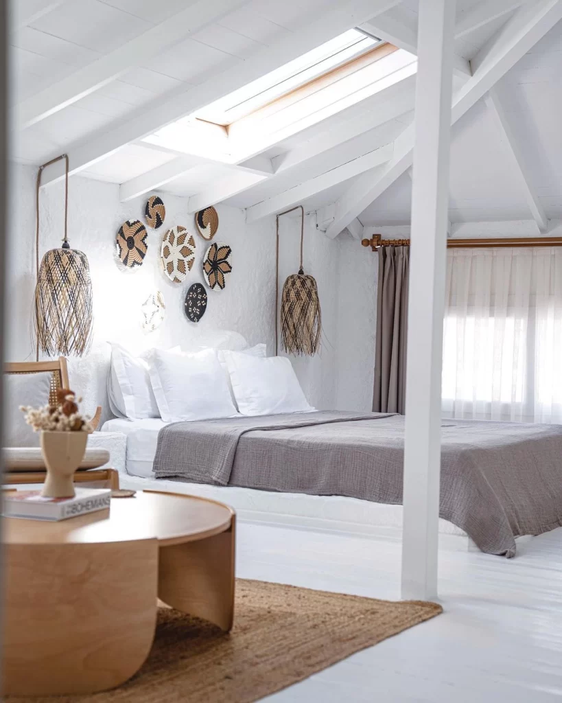 Beyaz ağırlıklı, eğimli tavanlı ve bol ışık alan tavan pencereli modern bir otel odasında, bembeyaz çarşaflarla kaplı ve açık gri pikeli yatağın başucunda, duvarda farklı desenlerde hasır duvar süslemeleri ve her iki yanında doğal dokulu sarkıt lambalar yer alıyor; odanın dingin ve şık atmosferini tamamlayan ahşap sehpa ve doğal jüt kilim ön planda dikkat çekiyor.
