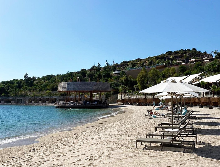 Bodrum'un en iyi otellerinden Le Meridien Bodrum'a ait geniş kum plajında, berrak turkuaz denizde yüzenlerin ve şezlonglarda beyaz şemsiyelerin gölgesinde dinlenenlerin olduğu sakin bir gün; arka planda yemyeşil tepelere yayılan modern otel binaları ve denizin ortasında ahşap çatılı bir tesis göze çarpıyor.