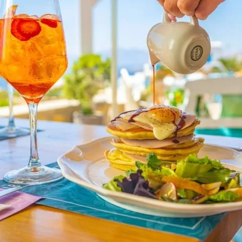 Mikonos'un eşsiz Ege manzarasına karşı, üzerinde kızarmış yumurta, jambon dilimleri ve sos gezdirilmiş kabarık pankeklerin yanında taze yeşilliklerle bezenmiş bir salata ve çilek dilimli serinletici turuncu bir içecek içeren, güne keyifli bir başlangıç vadeden zengin bir kahvaltı tabağı.