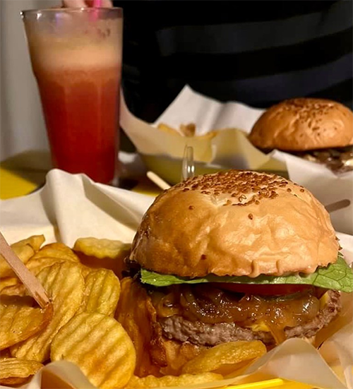 Susamlı ekmeği, erimiş peyniri, karamelize soğanları, marulu ve sulu köftesiyle iştah açıcı bir burgerin, çıtır tırtıklı patates cipsleriyle birlikte sunulduğu yakın çekim görüntüsü, Bodrum'un en iyi restoranlarından birinde unutulmaz bir lezzet deneyimine davet ediyor.