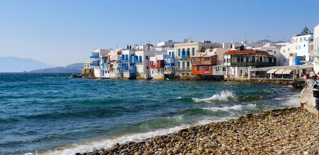 Ön plandaki çakıllı plaja vuran köpüklü dalgalarla canlanan masmavi Ege Denizi'ne sıfır, Mikonos'un meşhur Küçük Venedik bölgesindeki mavi, kırmızı ve kahverengi balkonlara sahip bembeyaz evler, adanın eşsiz güzelliğini güneşli bir havada gözler önüne seriyor.