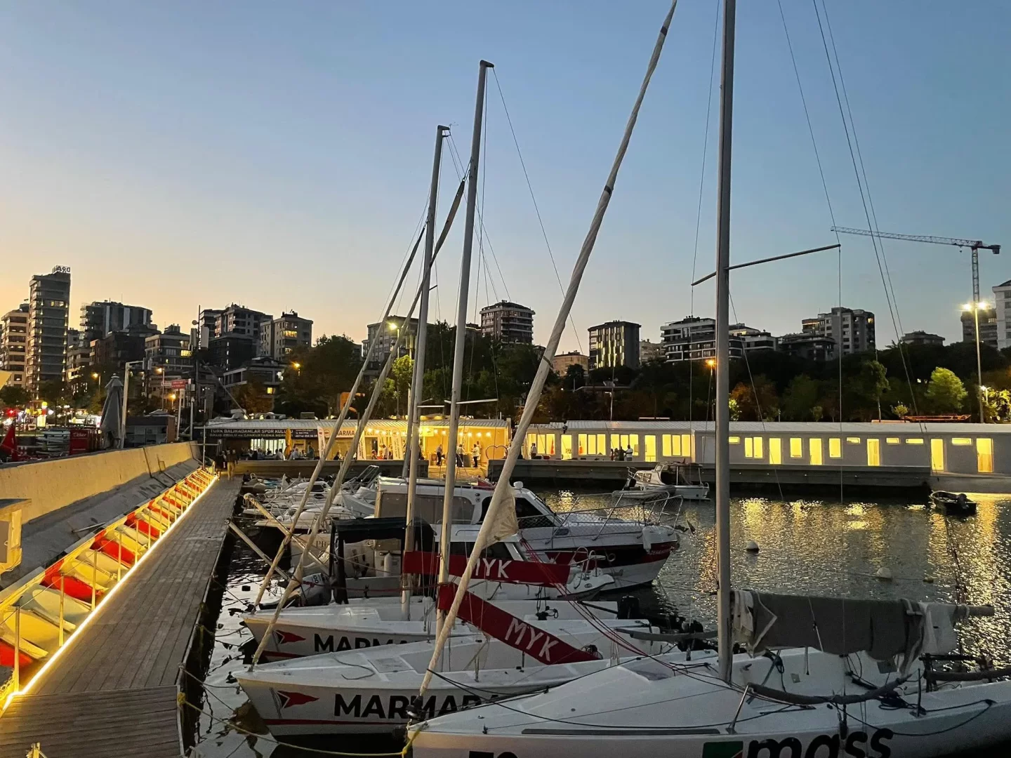 Alacakaranlıkta ışıl ışıl parlayan Marmara Yelken Kulübü marinasında, ahşap iskeleye bağlı bembeyaz yelkenliler, arkalarındaki modern şehir silüeti ve sudaki ışıltılarla huzurlu bir akşamüstü manzarası sunuyor.