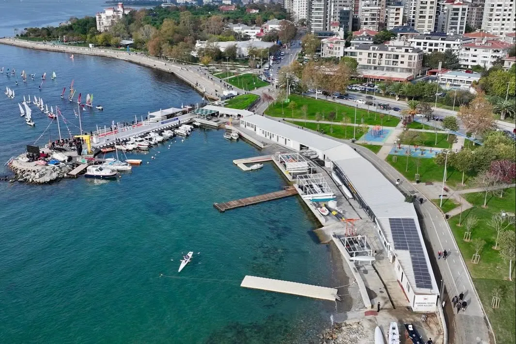 Marmara Yelken Kulübü'nün kuşbakışı görüntüsünde, deniz üzerinde çok sayıda yelkenlinin ve bir kayığın olduğu canlı bir aktivite alanı, solar panellerle kaplı uzun bir kulüp binası, rıhtımlar ve demirli tekneler ile kıyı şeridinde uzanan yeşil parklar, yürüyüş yolları ve şehir manzarası görülüyor.