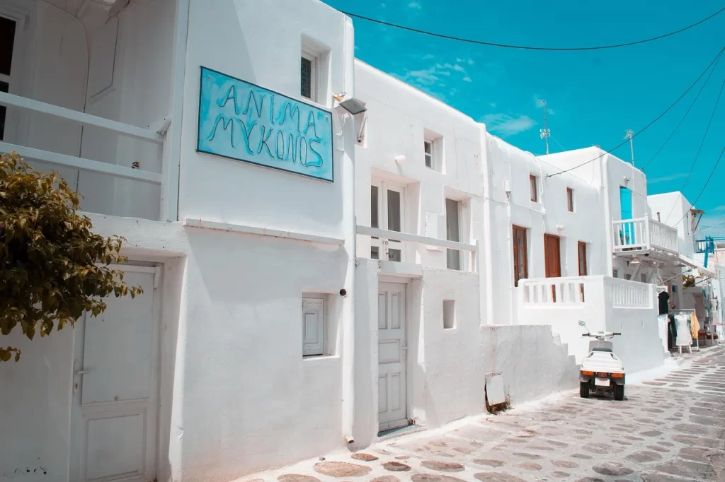 Mikonos'un kalbinde yer alan, bembeyaz badanalı evleri, dar ve taş döşeli sokağında park etmiş beyaz bir scooter ve parlak mavi gökyüzü ile adanın karakteristik mimarisini sergileyen bu görünüm, Mikonos seyahat rehberi içeriğindeki ruhu yansıtıyor.