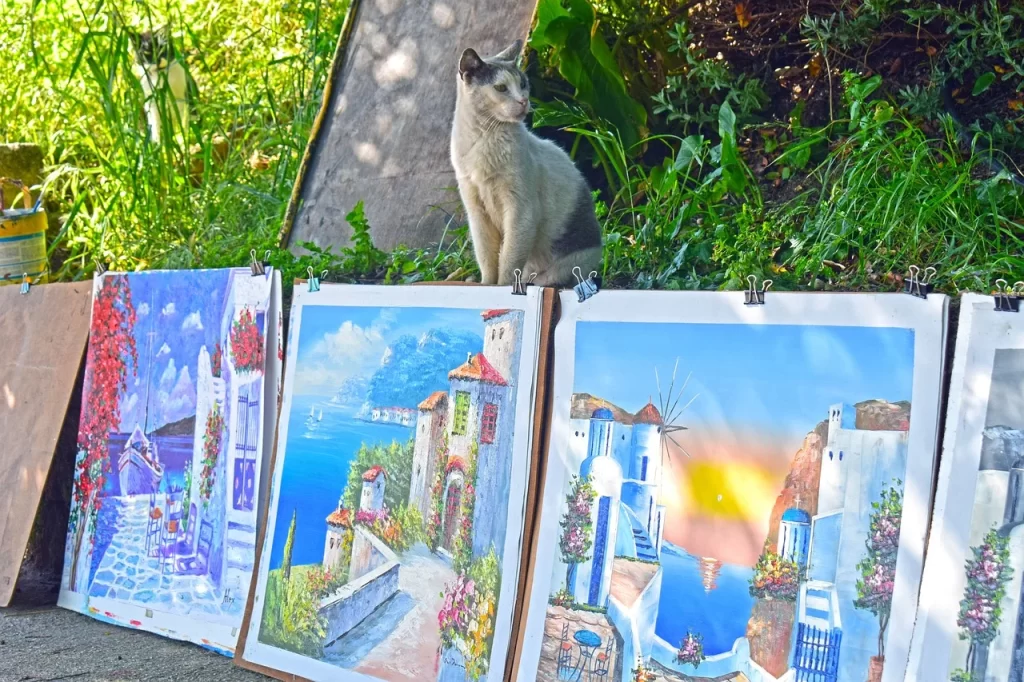 Bir kedi, Mikonos'a özgü bembeyaz evleri, yel değirmenlerini ve Ege Denizi'ni betimleyen renkli tabloların üzerinde otururken, adanın sanatçı ve tasarımcılarından Mikonos seyahati için kalıcı bir hatıra olabilecek özgün eserleri gözler önüne seriyor.