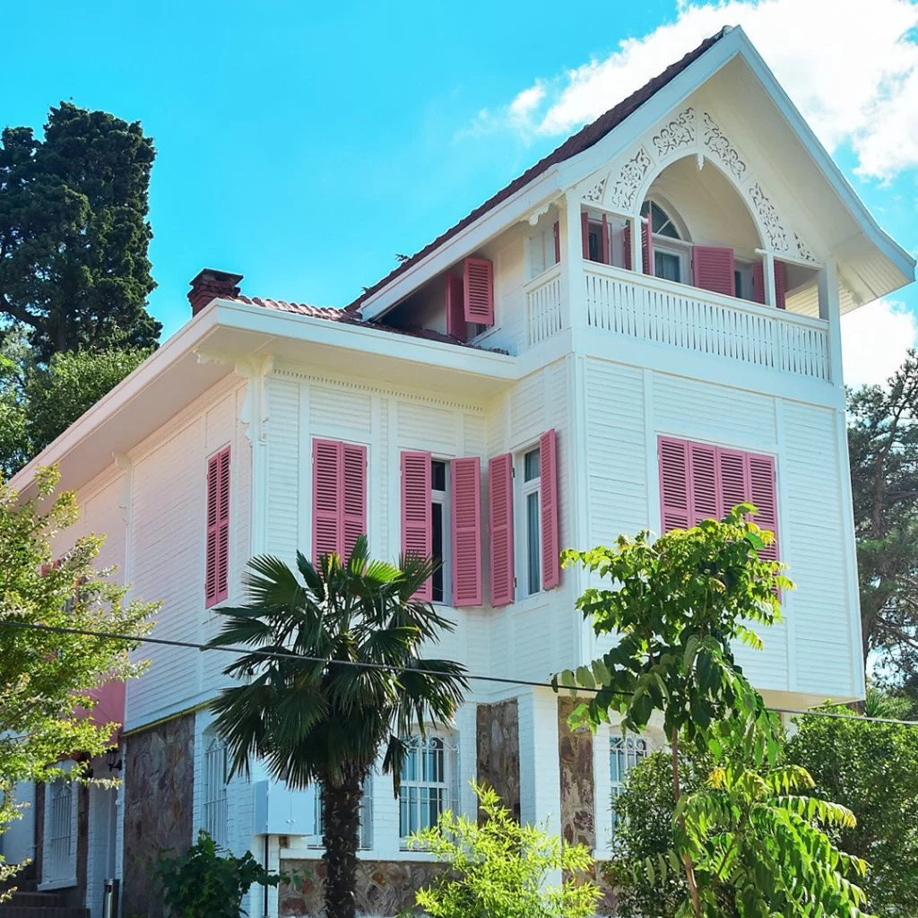 Mavi gökyüzü ve yemyeşil ağaçlar arasında yükselen, alt katı taş duvarlarla çevrili, beyaz ahşap cepheli, pastel pembe panjurlarıyla dikkat çeken ve üst katında oymalı ahşap korkuluklu bir balkon bulunan bu zarif bina, eski bir konak veya butik otel atmosferi sunuyor.