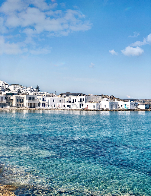 Paros adasının parlak güneşli bir gününde, berrak turkuaz denizin kıyısında sıralanmış, beyaz badanalı evleriyle ünlü, Ege mimarisinin güzelliğini yansıtan pitoresk bir sahil kasabasının manzarası.