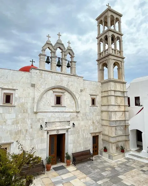 Bulutlu gökyüzü altında, açık renk mermer kaplı Panagia Tourliani Manastırı, üç çan kemeri ve yüksek, dört katlı bir çan kulesiyle Mikonos seyahati için önemli bir tarihi durak niteliğinde.