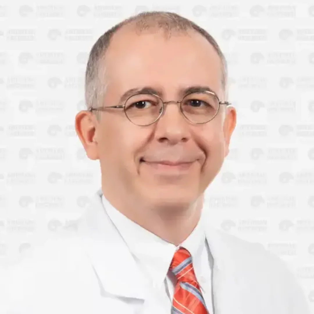Prof. Dr. Sinan Mahir Kayıran, açık tenli, yuvarlak gözlüklü, hafifçe seyrelmiş gri saçlı, beyaz önlüğü ve kırmızı-beyaz-mavi çizgili kravatıyla kameraya dostça gülümserken görünüyor; arka planda hafif bir hastane logosu deseni mevcut.