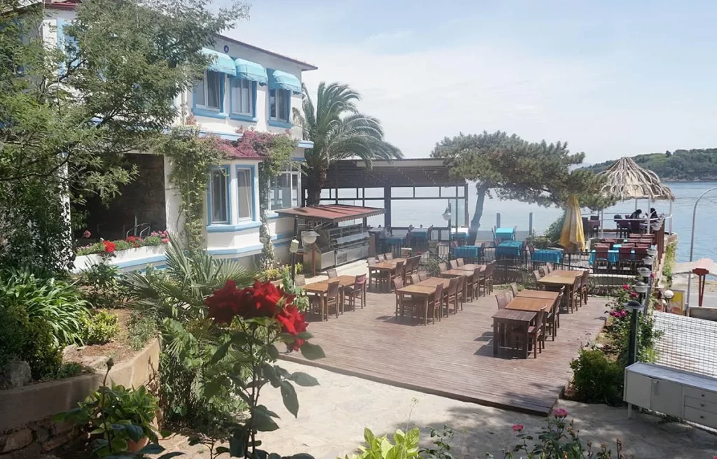 Burgazada'daki Pyrgos Otel'in beyaz duvarlı, mavi pencereli ve tenteli binasının önünde, kırmızı güllerle süslü yemyeşil bir bahçe uzanırken, deniz manzaralı geniş ahşap teras üzerindeki açık hava restoranının çok sayıda masası misafirlerini bekliyor.