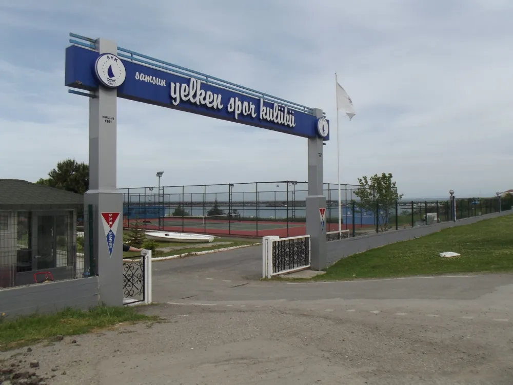 Samsun Yelken Spor Kulübü yazılı, mavi ve gri renklerdeki anıtsal giriş kemerinin önünden, kulübün kortlarını, uzakta görünen deniz manzarasını, çimlerin üzerinde duran bir yelkenli tekneyi ve solda küçük bir kulübeyi kapsayan genel bir görünüm.