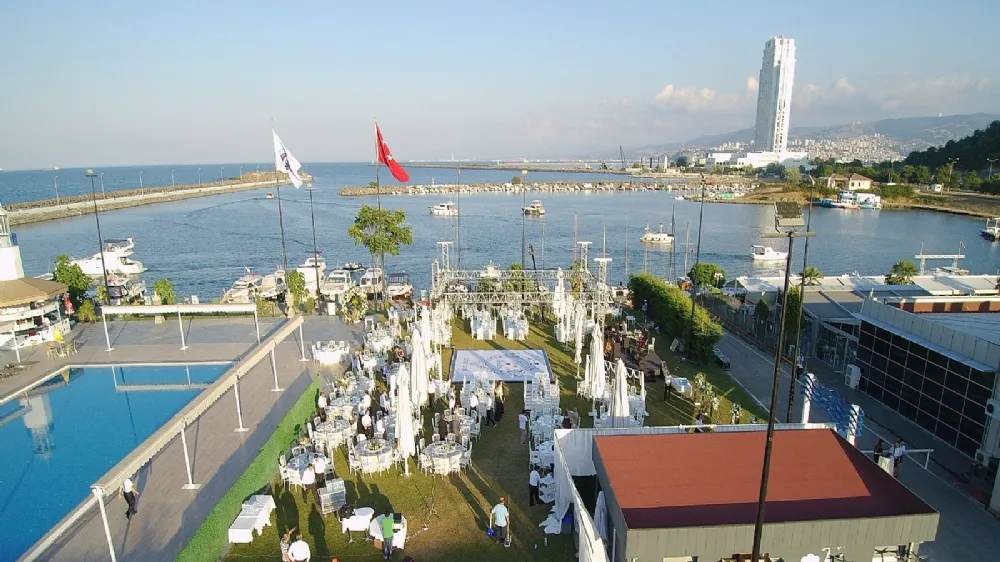 Samsun Yelken Kulübü'nün deniz manzaralı geniş yeşil alanında, beyaz masa ve sandalyelerle düzenlenmiş açık hava etkinliğinde misafirler sosyalleşirken, arka planda marinadaki tekneler ve şehrin simge yapısı modern bir gökdelen belirgin bir şekilde görülüyor.