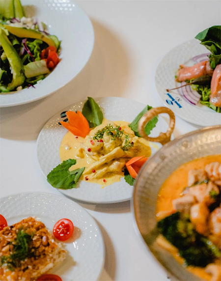 Beyaz bir masa üzerinde, ortasında kremalı soslu ve yeşilliklerle süslenmiş bir deniz ürünü ana yemeği ile birlikte, taze yeşil salata, karidesli meze, pirinç pilavı ve deniz mahsulleri çorbasının bol çeşitli bir serpme sunumu görülüyor.