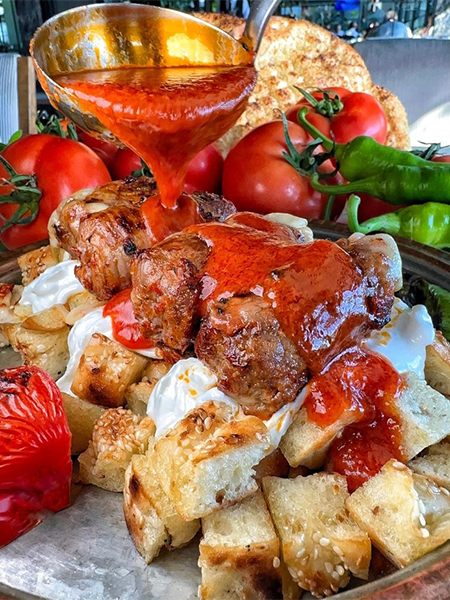 Metal bir tepside, kızarmış ekmek küpleri ve yoğurt üzerine dizilmiş ızgara kebap parçalarının parlak kırmızı bir sosla, arkada taze domatesler, yeşil biberler ve pide ekmeği eşliğinde sunulduğu iştah açıcı görüntü, Antalya’nın en iyi restoranlarında bulabileceğiniz geleneksel Türk kebap lezzetlerini vurguluyor.