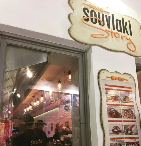 Mikonos adasının popüler restoranlarından Souvlaki Story’nin ön cephesini gösteren görselde, büyük bir tabela ve menü panosu dikkat çekerken, camdan yansıyan içerideki hareketlilik ve asılı dekoratif lambalar Ege mutfağının sıcak atmosferini yansıtıyor.