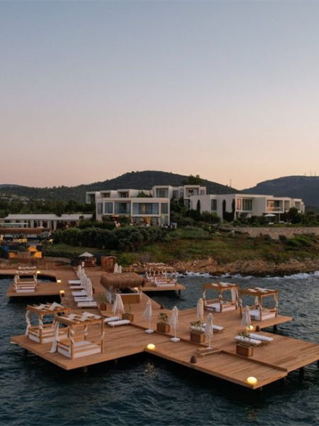 Alacakaranlıkta parlayan ahşap iskele, üzerindeki konforlu beyaz şezlonglar ve hasır çatılı localarla sakin denize uzanırken, yeşil yamaçlardaki modern beyaz otel binaları, Bodrum'un en iyi otellerinden birinin sofistike ve huzurlu atmosferini gözler önüne seriyor.