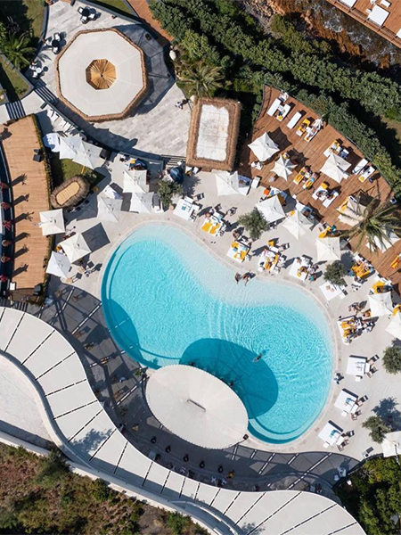 Bodrum'un en iyi otellerinden birinde, berrak mavi havuzun çevresine yerleştirilmiş beyaz şezlong ve şemsiyelerle dolu, yeşil bitki örtüsüyle çevrili lüks bir tatil köyünün kuşbakışı görüntüsü, huzurlu bir dinlenme ortamı sunuyor.