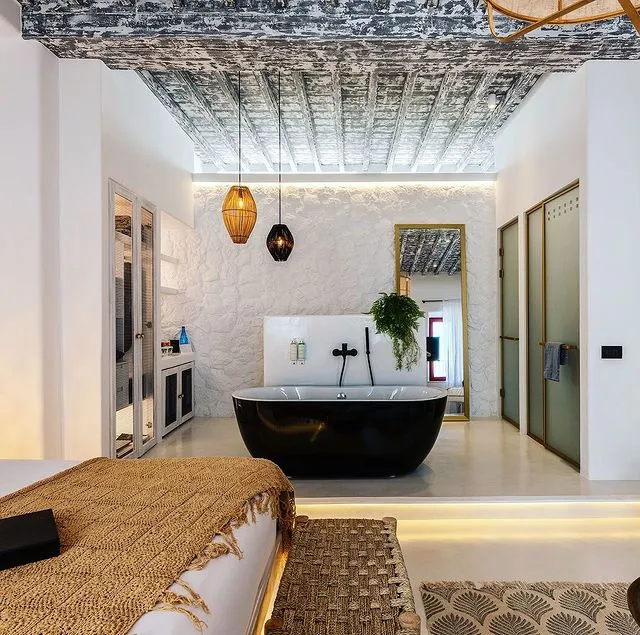 Mikonos'ta unutulmaz bir seyahat deneyimi vadeden, beyaz dokulu duvarlara, rustik ahşap tavan kirişlerine, şık siyah bir küvete ve modern tasarımlı aydınlatmalara sahip ferah ve lüks bir otel süitinin iç görünümü.