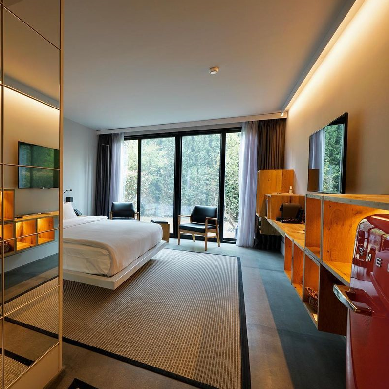 Modern bir otel odasında, aynalı bir duvar, beyaz nevresimli geniş bir yatak, orman manzarasına açılan boydan boya pencereler, ahşap raflı medya ünitesi, duvara monte TV ve kırmızı buzdolabı ile şık bir konaklama alanı sunuluyor.