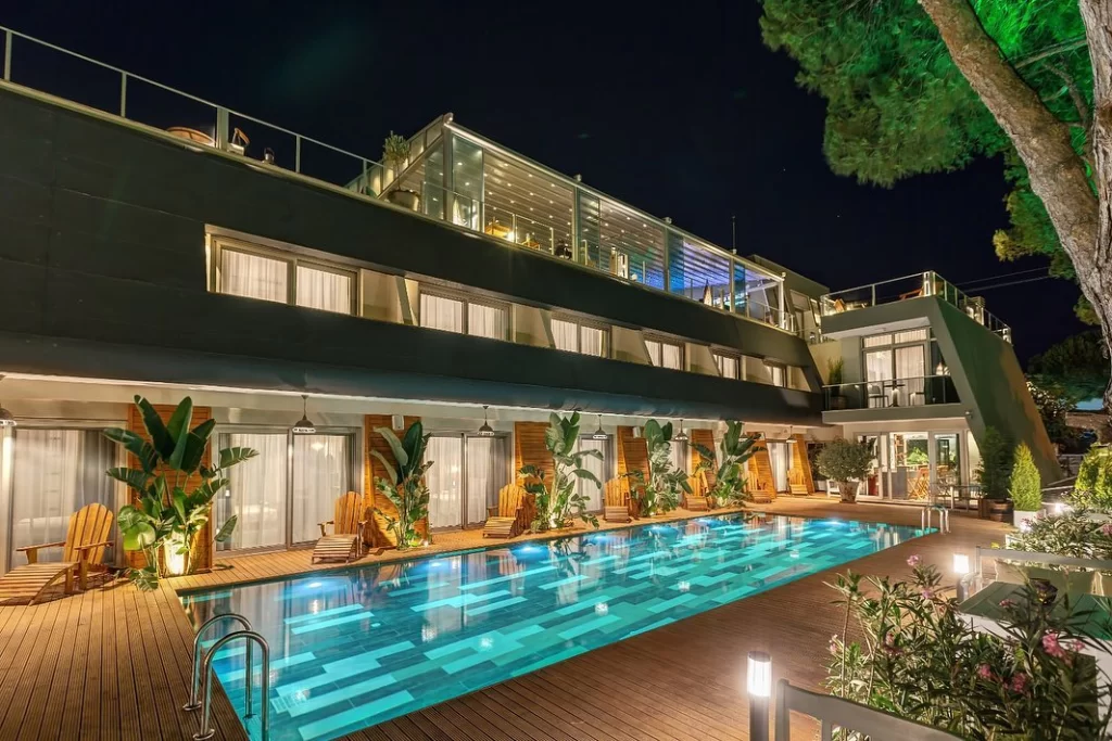 Modern bir villa veya otel, gece ışıklandırmasıyla aydınlanmış, ahşap bir zeminle çevrili, içinde mavi yansımaların olduğu uzun bir yüzme havuzu, havuz kenarında ahşap şezlonglar ve tropikal bitkilerle birlikte görünüyor; binanın üst katında cam panelli bir çatı terası yer alıyor ve arka planda yeşil ışıklarla aydınlatılmış büyük bir ağaç göze çarpıyor.