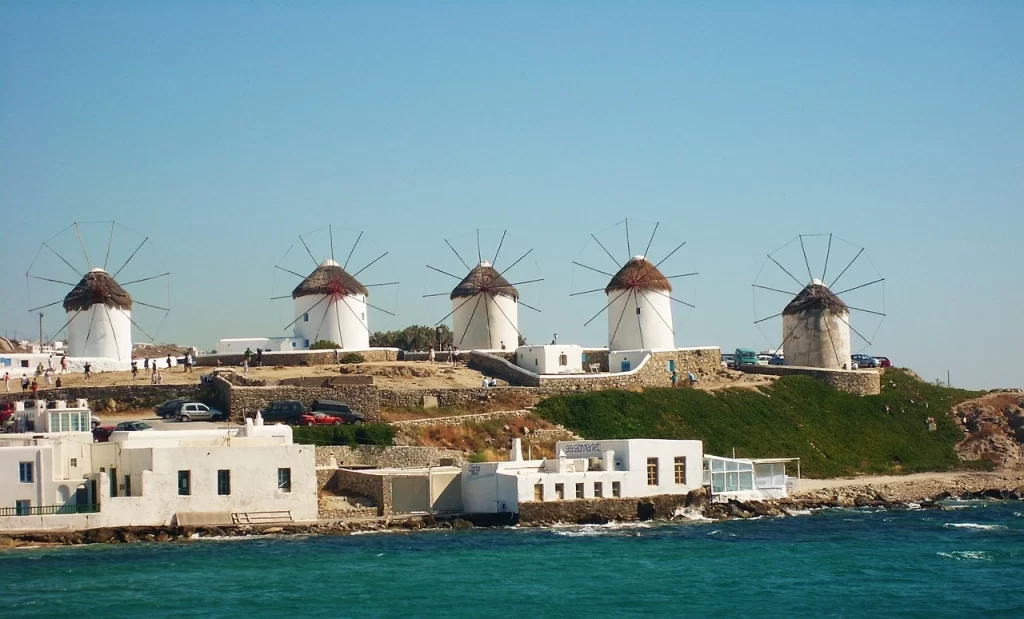 Mikonos adasının berrak Ege suları kıyısında yer alan, beyaz badanalı evlerin üzerinde sıralanmış beş ikonik yel değirmeni, adanın tarihi dokusunu ve doğal güzelliğini güneşli bir günde gözler önüne seriyor.