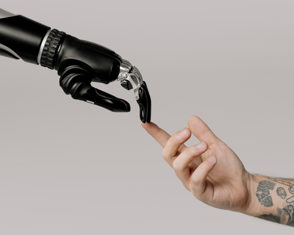 Açık gri bir fonda, modern teknolojinin simgesi olan siyah ve metalik detaylı bir robot elin, dövmeli bir insan elinin işaret parmağına nazikçe dokunduğu bu etkileyici görsel, yapay zekâ ve insan etkileşimi gibi konulara olan artan ilgiyi yansıtarak, sunduğumuz farklı ilgi alanlarına yönelik podcast önerileri listesinde özellikle teknoloji odaklı bölümlerin önemine işaret ediyor.