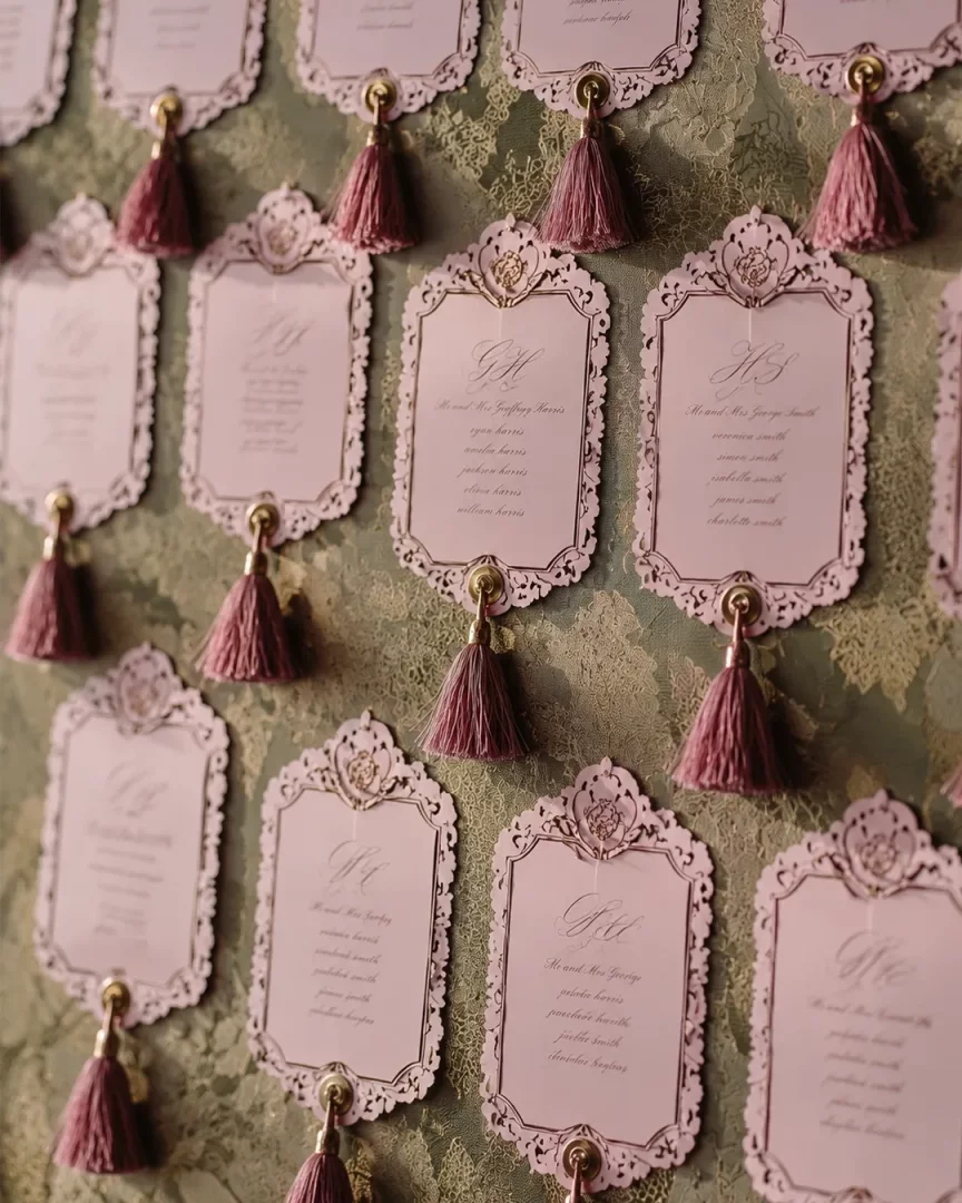 Yeşil damask desenli bir zemine asılmış, kenarları lazer kesim detaylı pembe renkli ve püsküllü zarif düğün davetiyeleri, kişiselleştirilmiş isim listeleriyle 2026 düğün trendlerinin yeni formda davetiyelerini yansıtıyor.
